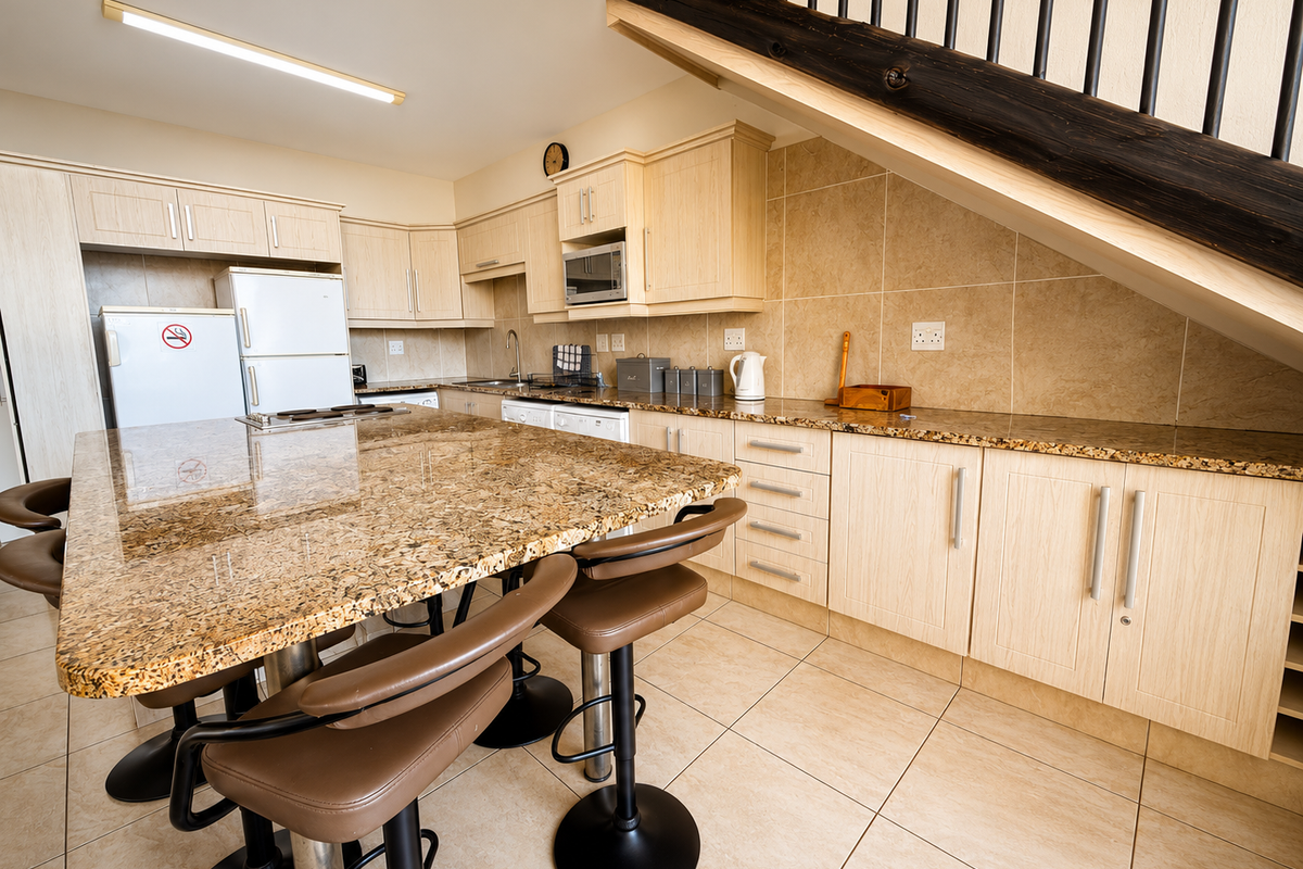 A functional kitchen featuring granite countertops, light wood cabinetry, built-in appliances, and tiled flooring, situated beneath a staircase.