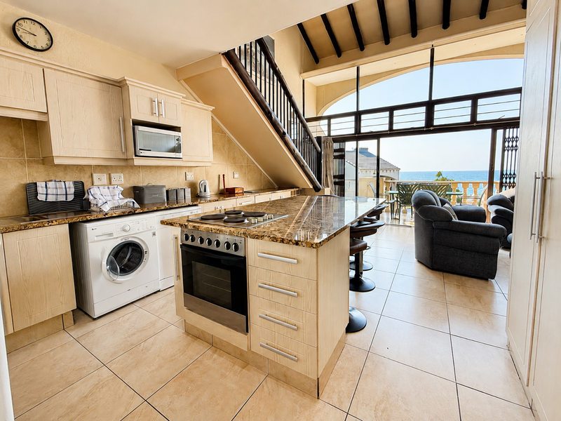 Spacious open-plan kitchen and living area featuring granite countertops, built-in appliances, tiled floors, and large sliding doors leading to a balcony with ocean views.