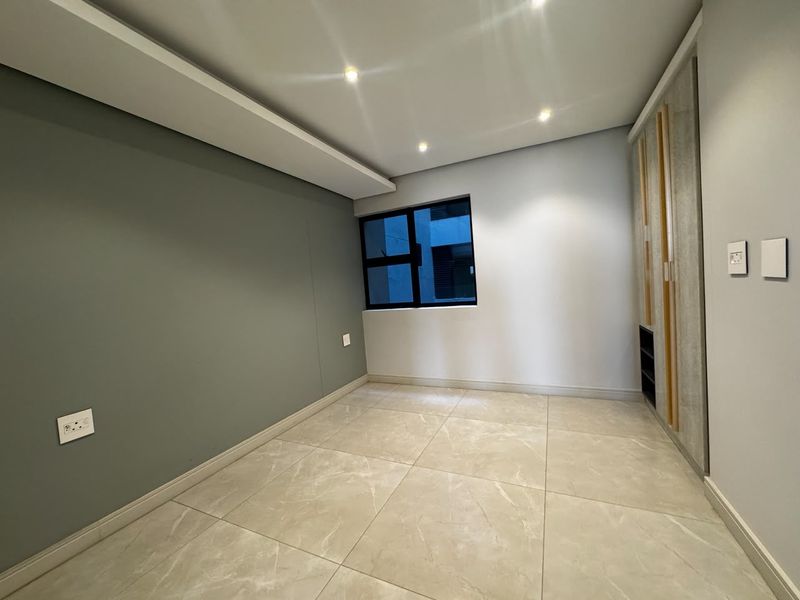 A modern, empty bedroom featuring neutral tiled flooring, built-in cupboards, a bulkhead ceiling with recessed lighting, and a large aluminium-framed window.
