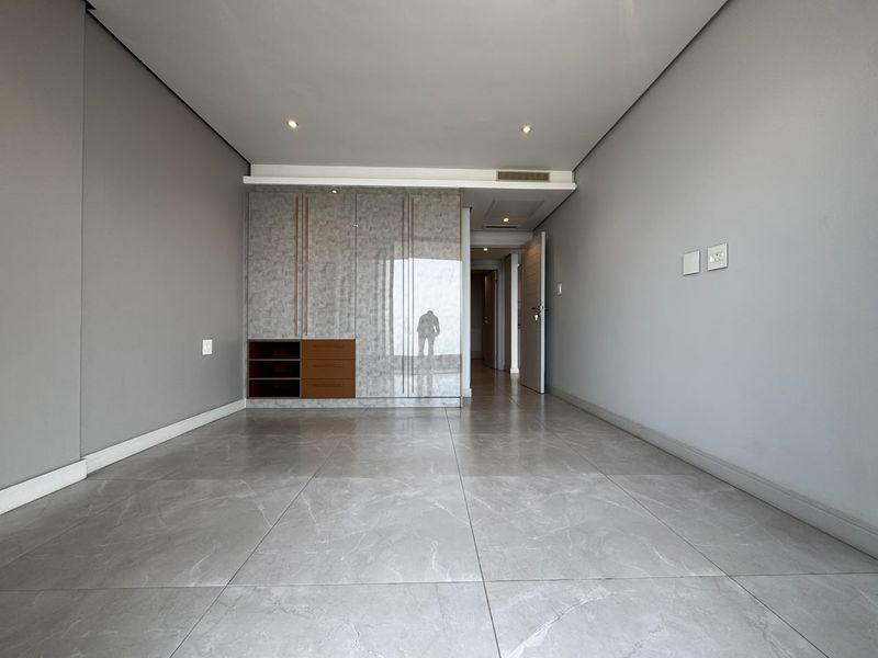 A modern, empty bedroom featuring large-format grey porcelain floor tiles, built-in cupboards with a glossy finish, and recessed ceiling lighting.