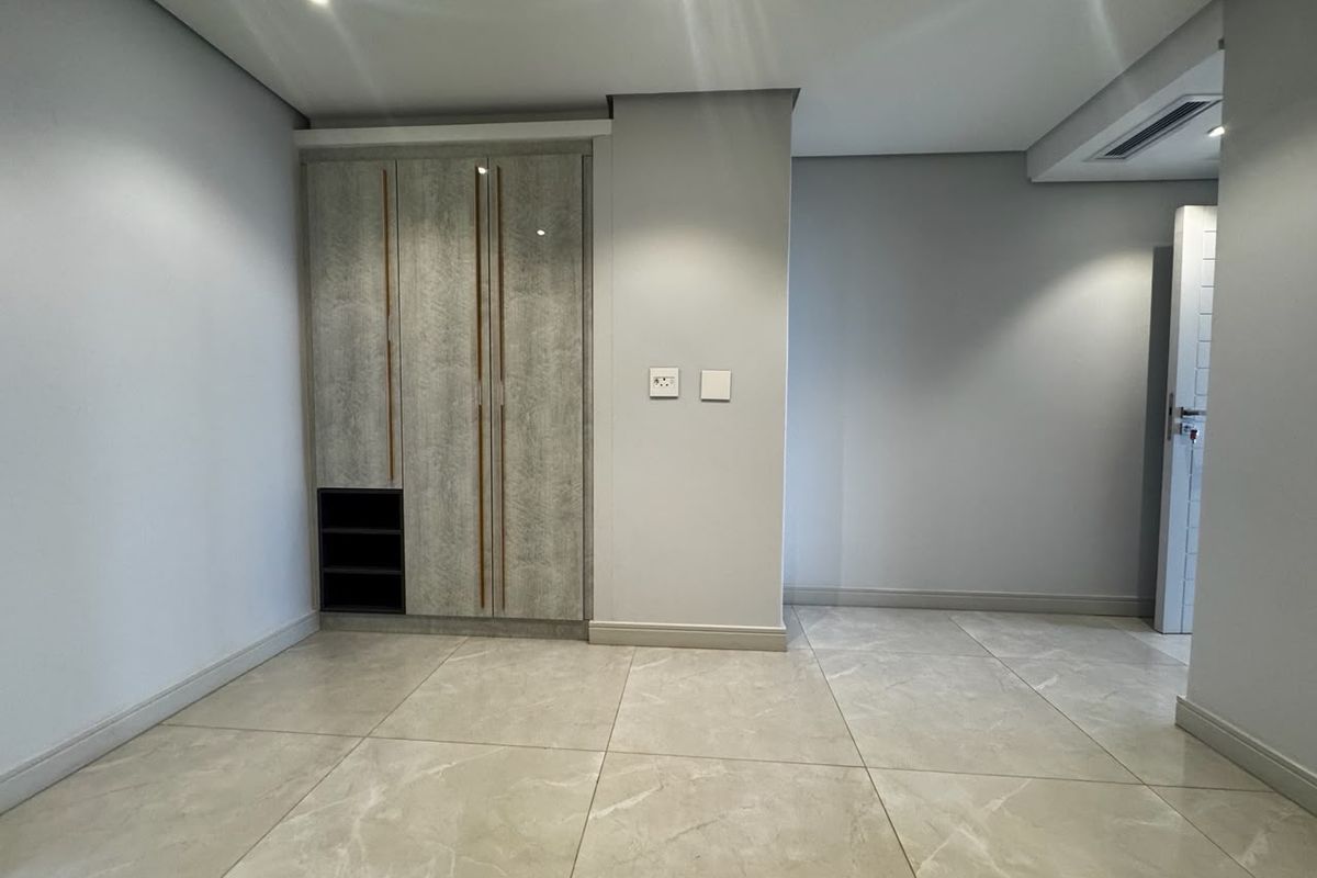 A clean, modern bedroom featuring neutral porcelain floor tiles, recessed ceiling lighting, and a contemporary built-in wardrobe with integrated shelving.