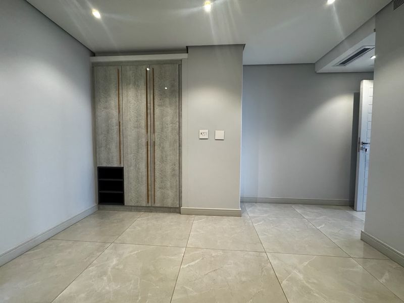 A clean, modern bedroom featuring neutral porcelain floor tiles, recessed ceiling lighting, and a contemporary built-in wardrobe with integrated shelving.