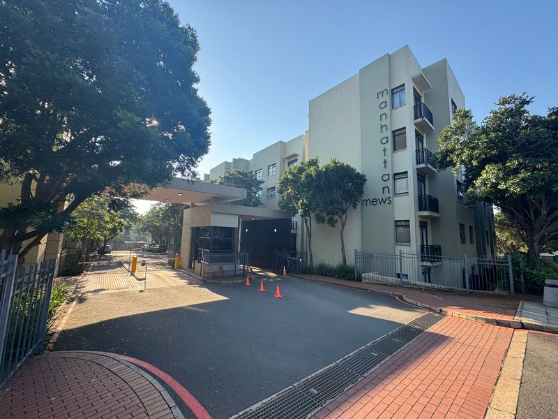 Front entrance view of Manhattan Mews apartment complex featuring a secure gatehouse, paved driveway, and multi-story building facade with balconies.