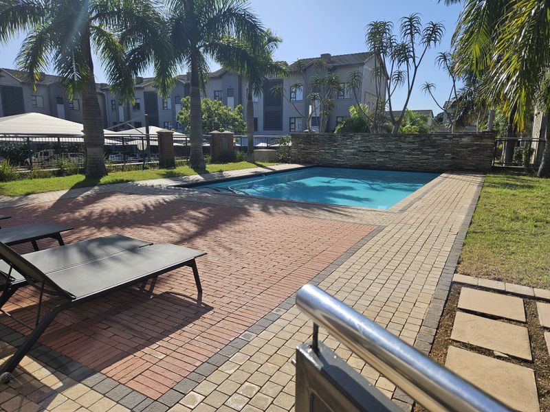 Communal swimming pool area featuring a stone feature wall, paved decking, and landscaped surroundings within the apartment complex.