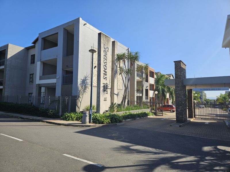 Exterior view of the Seasons Court apartment complex entrance, featuring modern facade architecture, secure gated access, and paved driveway.