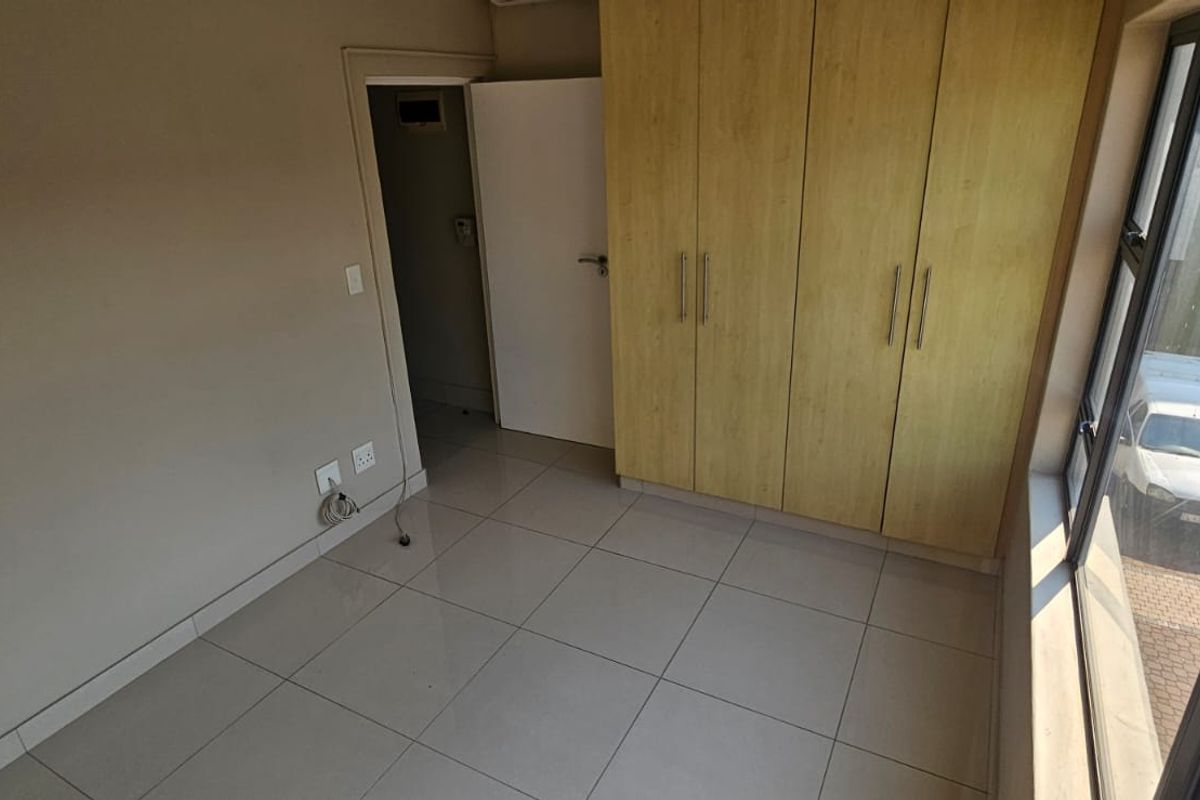 A bright, empty bedroom featuring neutral tiled flooring, built-in wooden cupboards, and large windows providing natural light.