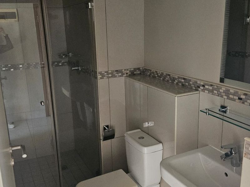 A clean, functional bathroom featuring a glass shower enclosure, wall-mounted toilet, and vanity basin with neutral tiling and mosaic accents.