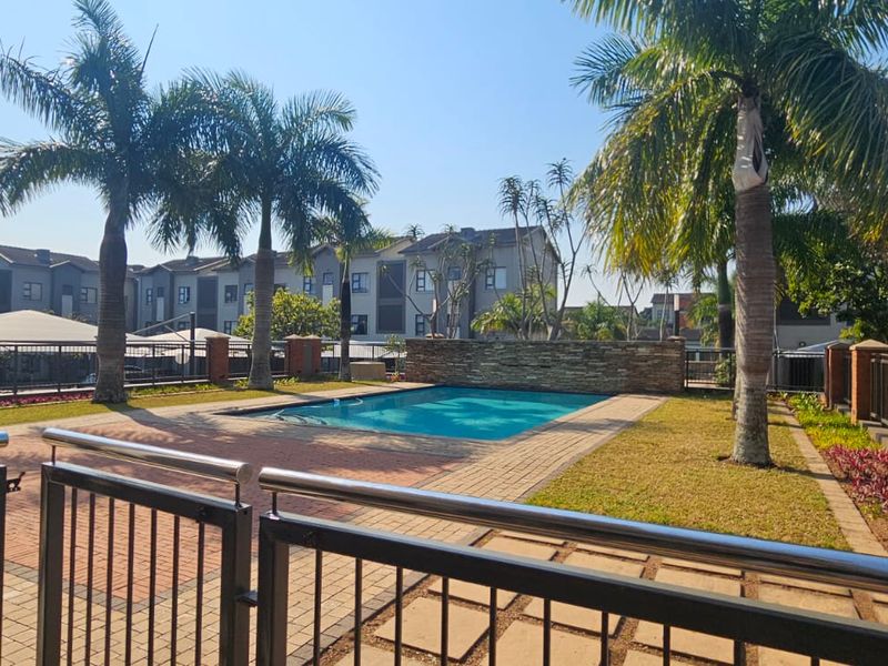 A well-maintained communal swimming pool area featuring brick paving, a stone feature wall, and established palm trees within a secure residential complex.
