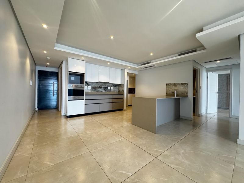 Modern open-plan kitchen and living area featuring sleek cabinetry, a stone island, recessed lighting, and large format floor tiles in a contemporary apartment setting.