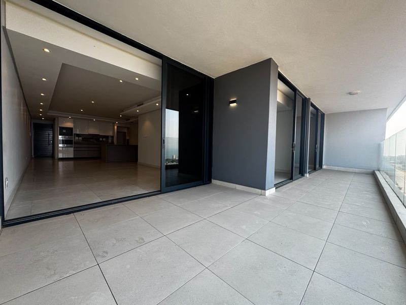 Spacious covered balcony featuring large format floor tiles, modern glass balustrades, and seamless access to the open-plan interior living area.