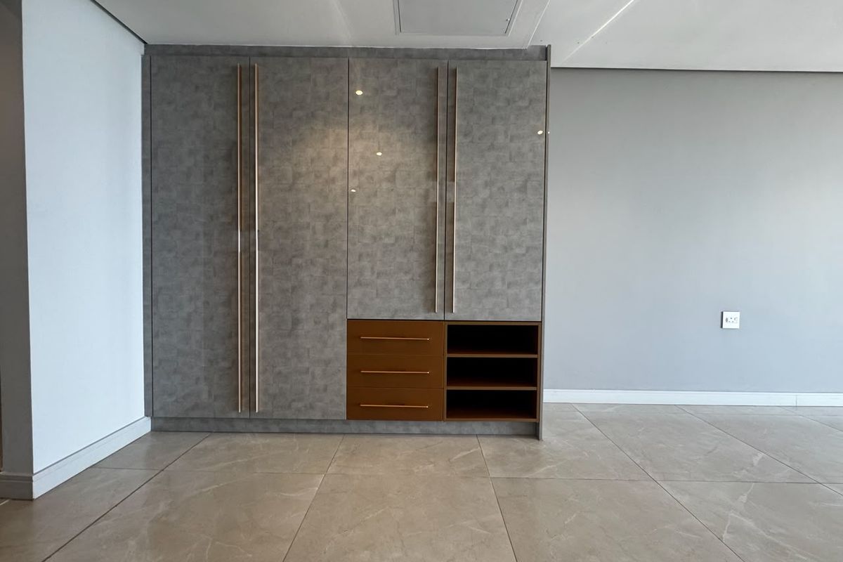 A modern, empty bedroom featuring sleek built-in cupboards with integrated drawers and shelving, complemented by neutral tiled flooring and recessed ceiling lighting.