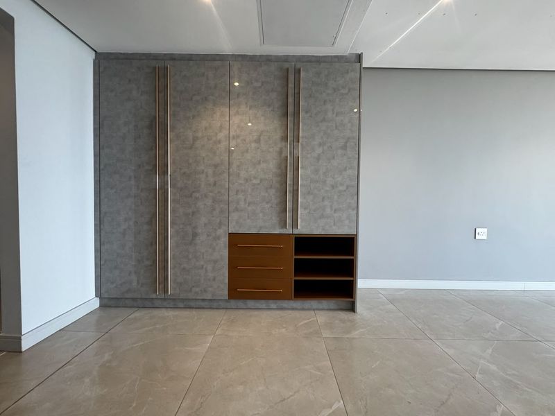 A modern, empty bedroom featuring sleek built-in cupboards with integrated drawers and shelving, complemented by neutral tiled flooring and recessed ceiling lighting.