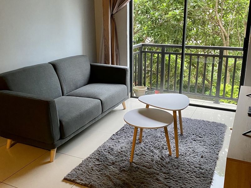 A bright living room featuring large sliding glass doors that open onto a balcony with lush greenery views and neutral tiled flooring.