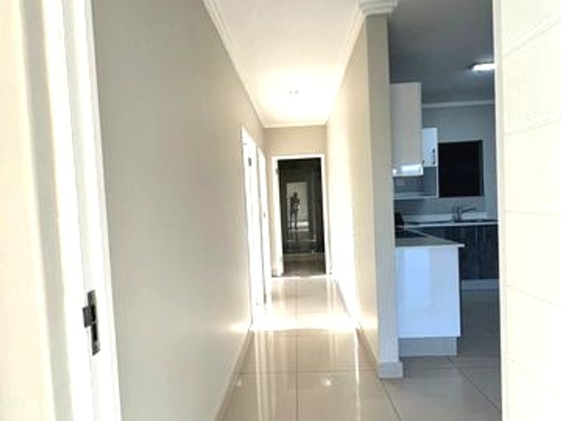 A bright, modern hallway featuring neutral walls, large format floor tiles, and recessed downlighting, leading towards an open-plan kitchen area.