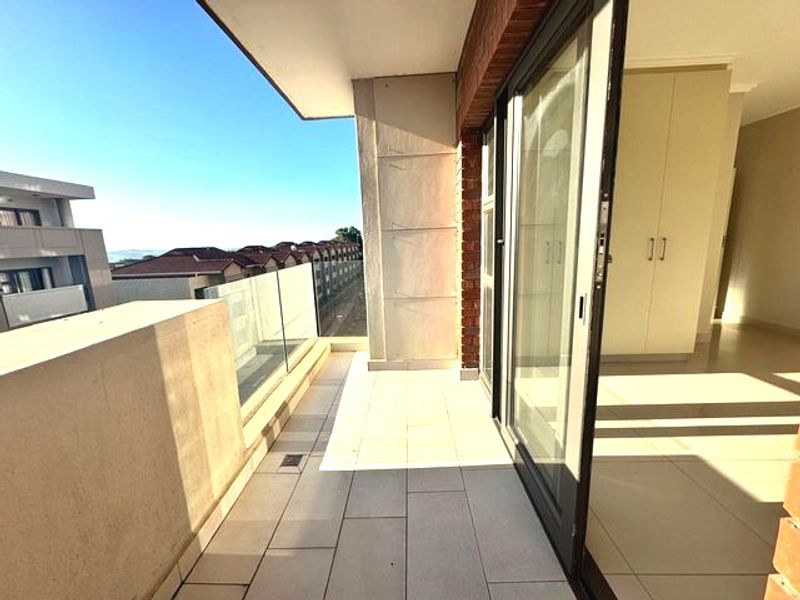 A bright, modern balcony featuring tiled flooring, a sleek glass balustrade, and direct access to the interior through large sliding glass doors.
