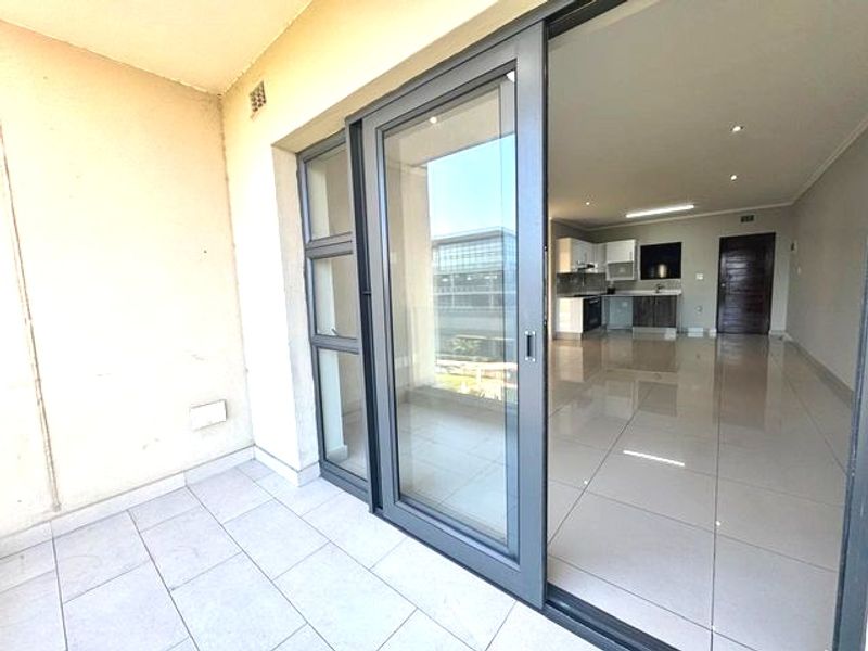 A covered balcony with tiled flooring, featuring a large sliding glass door leading into an open-plan living and kitchen area.