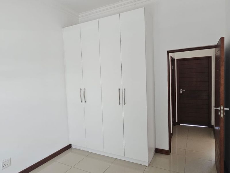 A clean, empty bedroom featuring white built-in cupboards, neutral tiled flooring, and a dark wood internal door leading to the hallway.