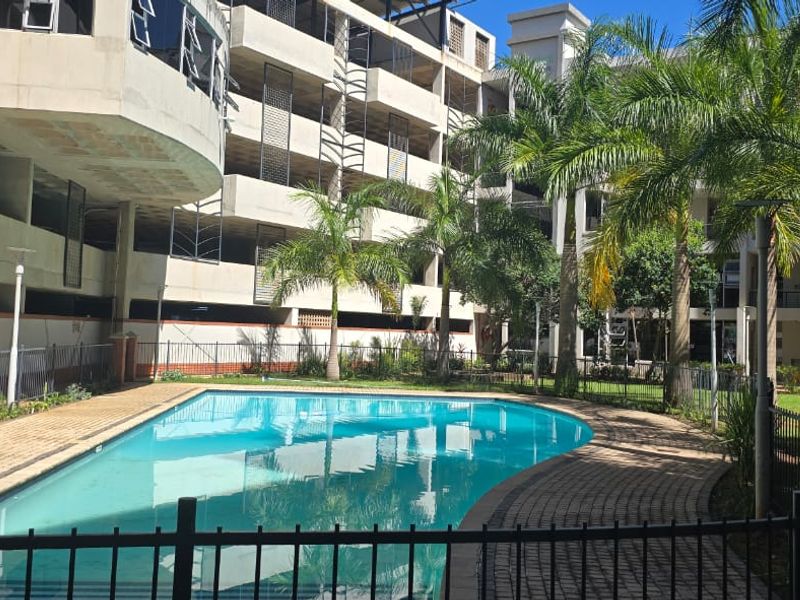 A well-maintained communal swimming pool area featuring paved surrounds, metal perimeter fencing, and established palm trees within an apartment complex setting.