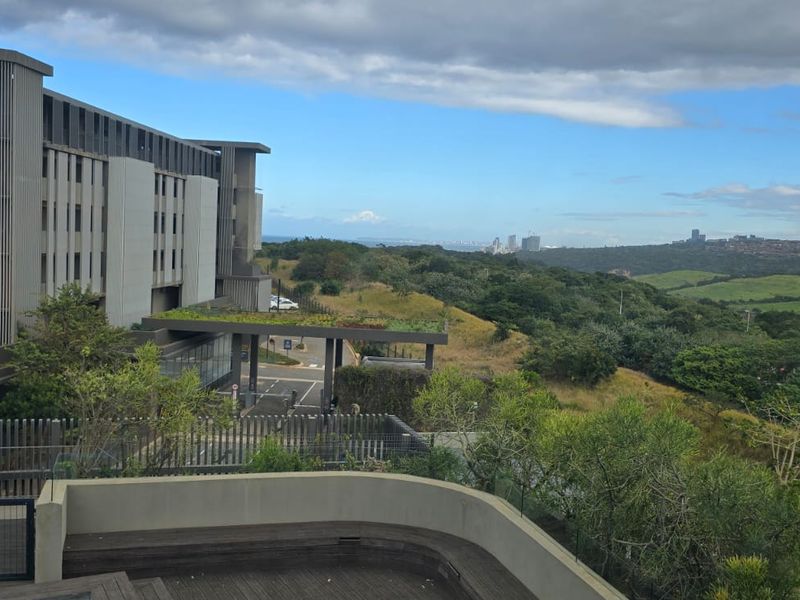 Elevated view overlooking the apartment complex, surrounding green belt, and distant city skyline, featuring a modern timber deck and glass balustrade.