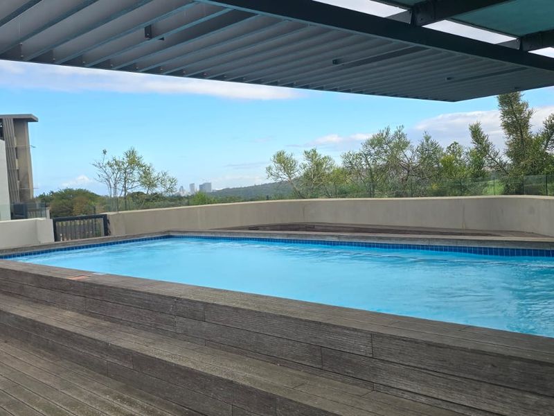 A modern raised swimming pool area featuring timber decking, a louvred pergola for shade, and a glass balustrade with scenic views.