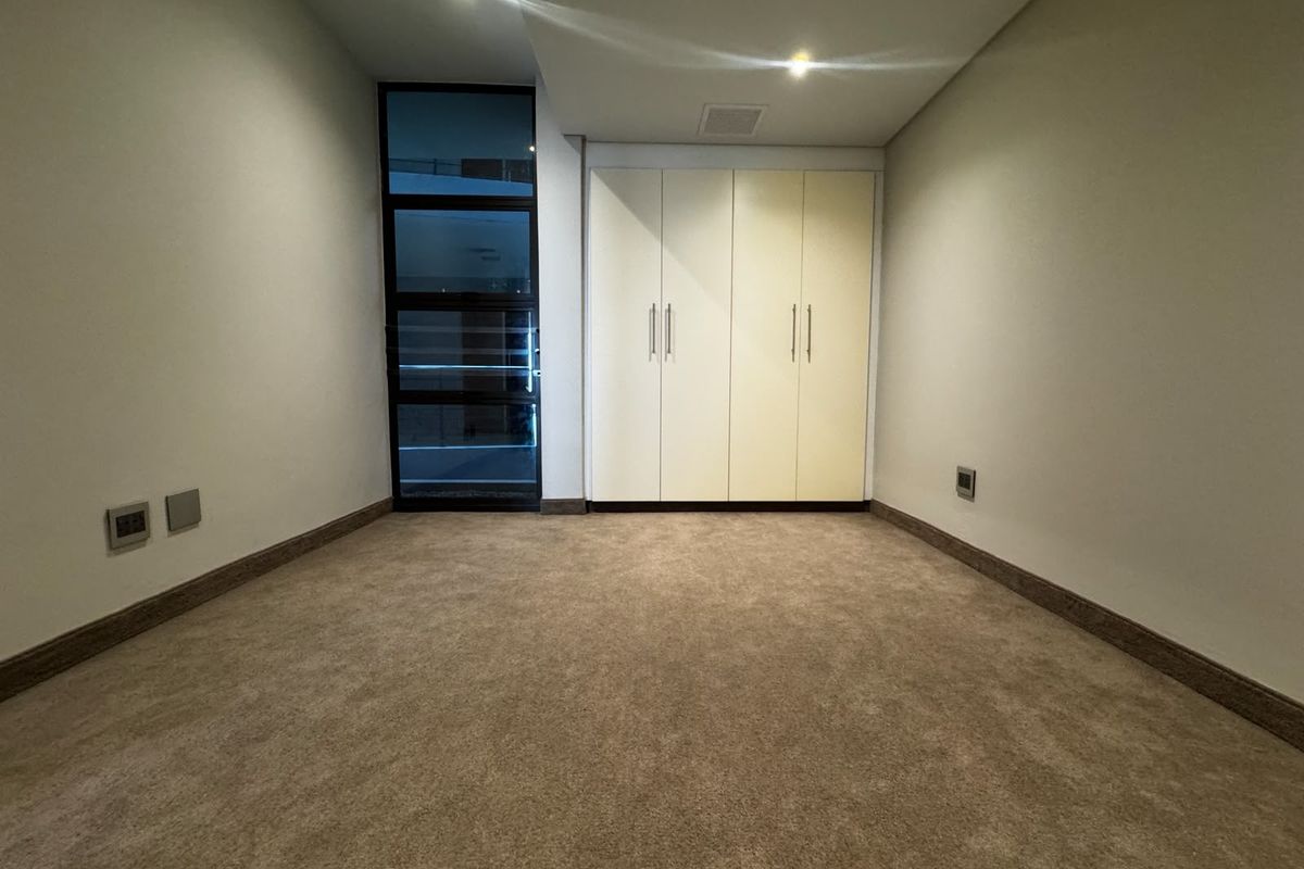 A clean, empty bedroom featuring neutral carpeting, built-in cupboards, and a modern glass-panelled door providing natural light.