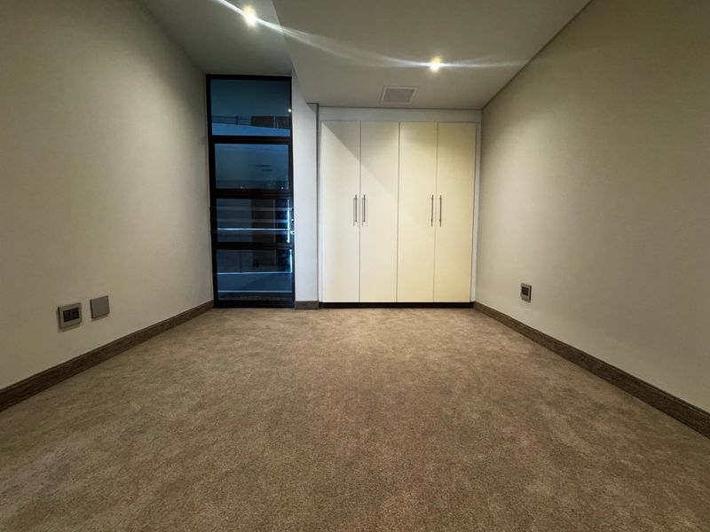 A clean, empty bedroom featuring neutral carpeting, built-in cupboards, and a modern glass-panelled door providing natural light.