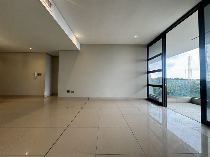 A bright, empty living area featuring large sliding glass doors that open onto a balcony with a glass balustrade and tiled flooring throughout.