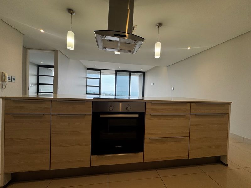 Modern open-plan kitchen featuring a central island with built-in oven, extractor fan, stone countertops, and sleek cabinetry, flowing into a bright living area.