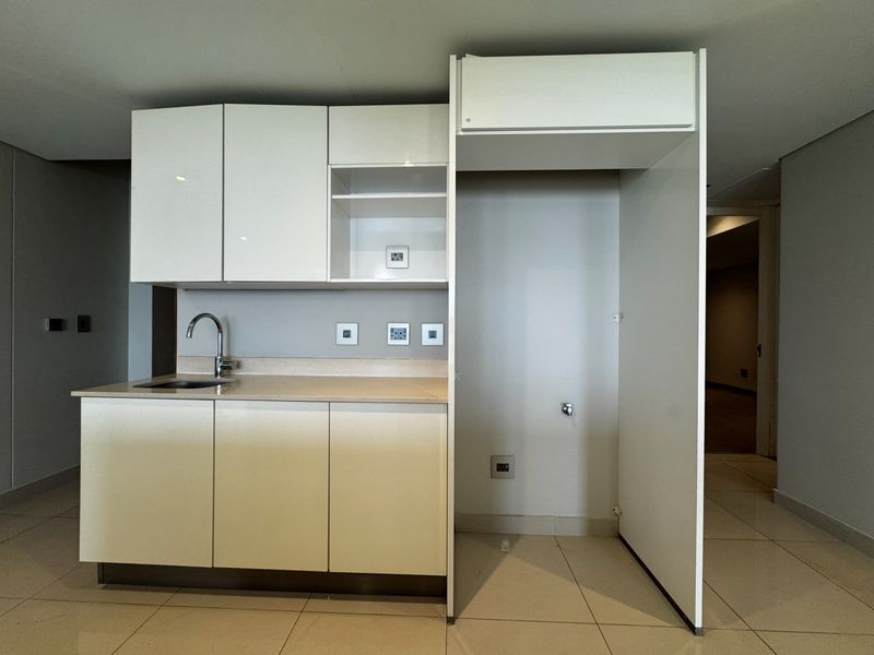 A clean, modern kitchen area featuring white built-in cabinetry, a stainless steel sink, and a dedicated alcove for a refrigerator.