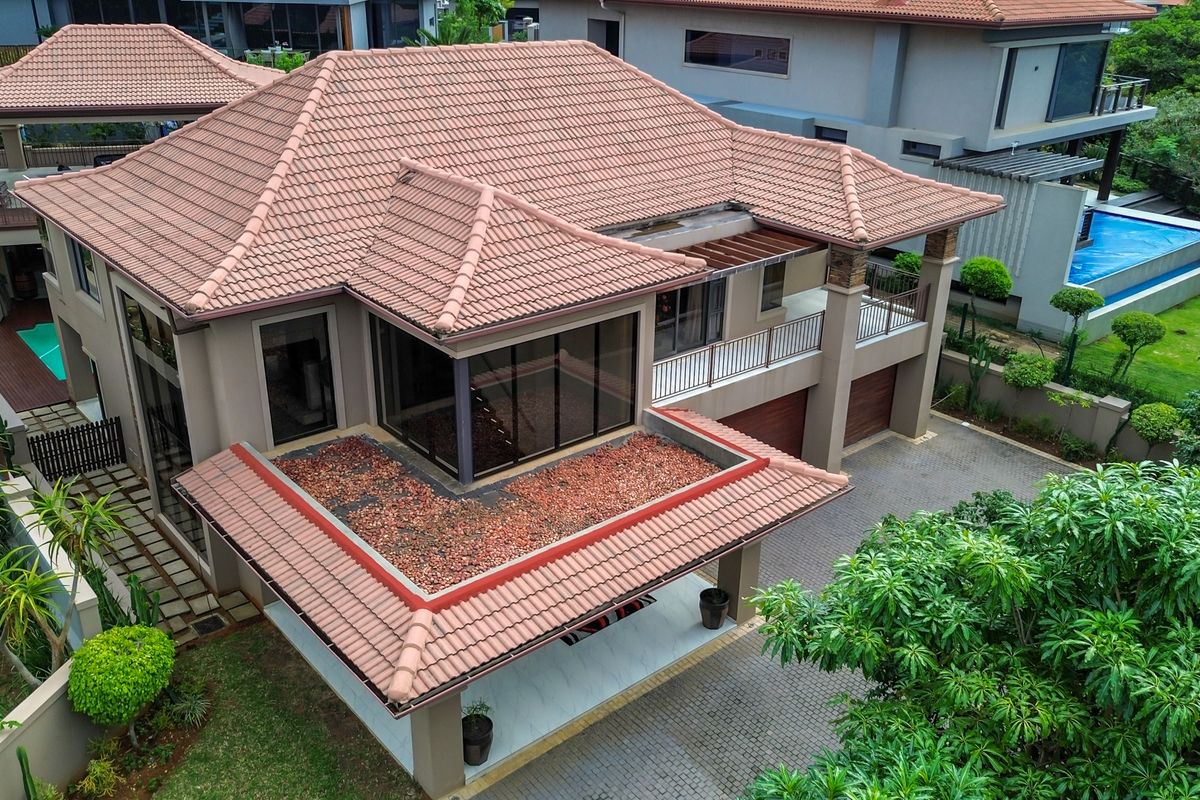 Aerial view of a modern residential property featuring a terracotta tiled roof, paved driveway, double garage, balcony, and a well-maintained garden with a pool.