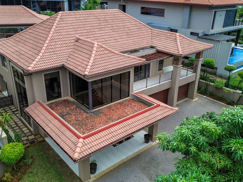 Aerial view of a modern residential property featuring a terracotta tiled roof, paved driveway, double garage, balcony, and a well-maintained garden with a pool.