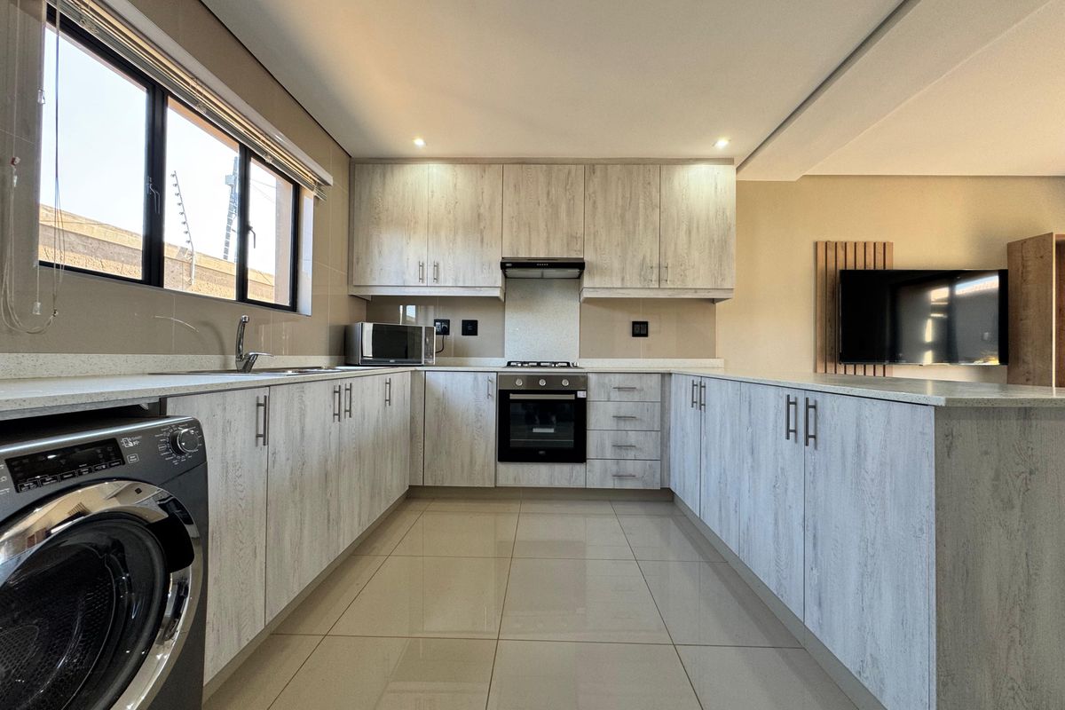 Modern kitchen featuring light wood-grain cabinetry, stone countertops, integrated oven and hob, and sleek tiled flooring in an open-plan layout.