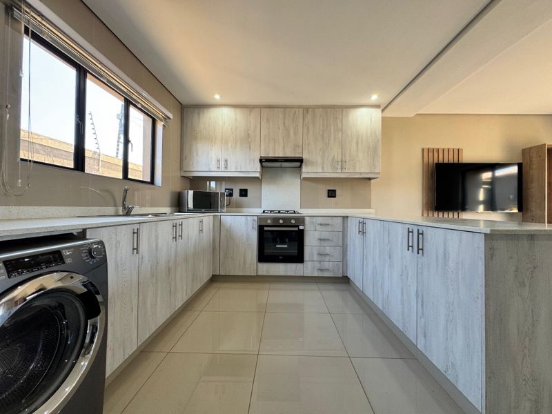 Modern kitchen featuring light wood-grain cabinetry, stone countertops, integrated oven and hob, and sleek tiled flooring in an open-plan layout.