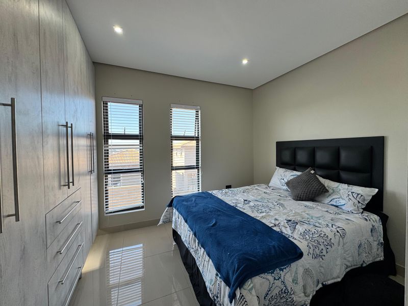 A modern bedroom featuring neutral tiled flooring, ample built-in cupboard storage, recessed lighting, and dual windows with fitted venetian blinds.