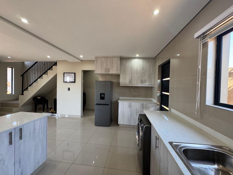 Modern open-plan kitchen area featuring light wood-grain cabinetry, stone countertops, tiled flooring, and an integrated staircase with a metal balustrade.
