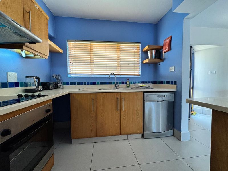 A functional kitchen featuring wooden cabinetry, tiled splashbacks, built-in oven, hob, extractor fan, and integrated dishwasher with tiled flooring.