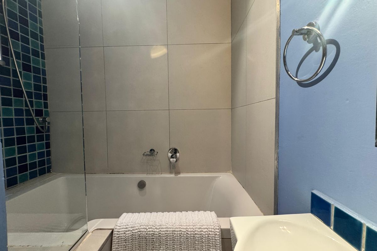 A functional bathroom featuring a bathtub with a glass shower screen, neutral wall tiling, and a blue accent wall with a vanity basin.