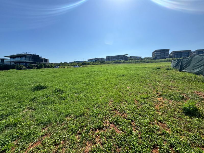 A wide-angle view of a vacant residential plot featuring open grass terrain and surrounding neighborhood development.