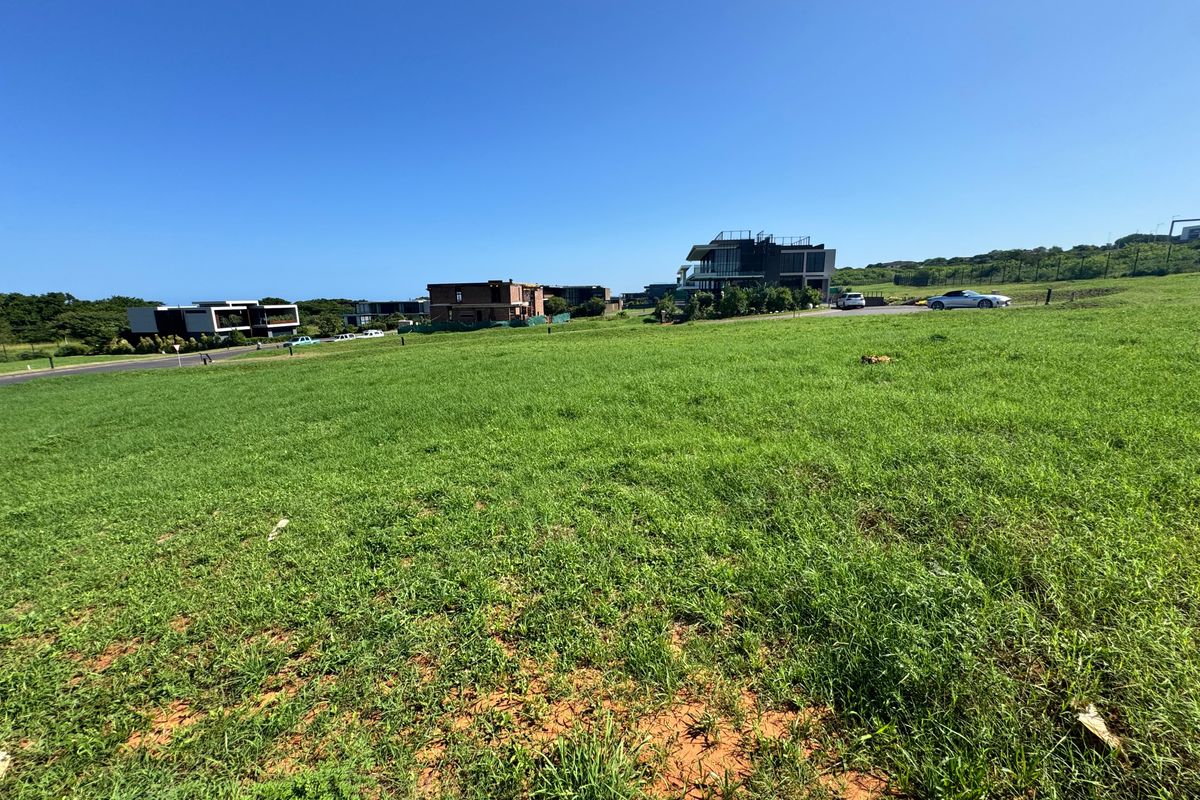 A vacant residential plot of land featuring open grass terrain with established neighbourhood infrastructure and road access in the surrounding area.
