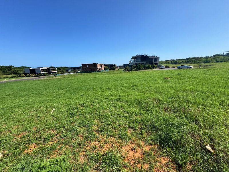 A vacant residential plot of land featuring open grass terrain with established neighbourhood infrastructure and road access in the surrounding area.