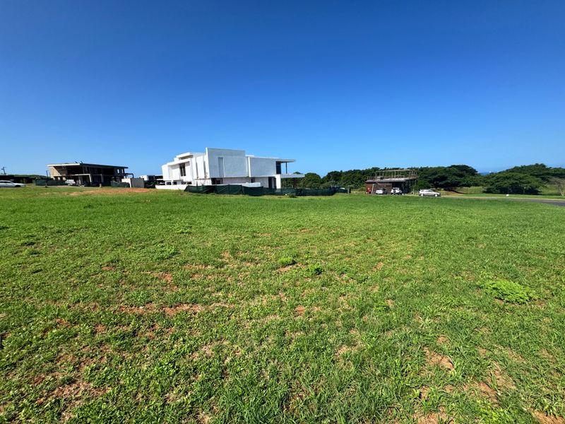 A wide-angle view of a vacant residential plot featuring a grassy landscape with neighboring modern properties visible in the background.