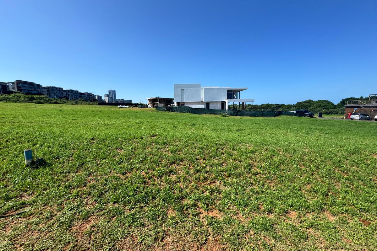 A wide-angle view of a vacant residential plot featuring open grassed land with surrounding neighborhood development visible in the background.