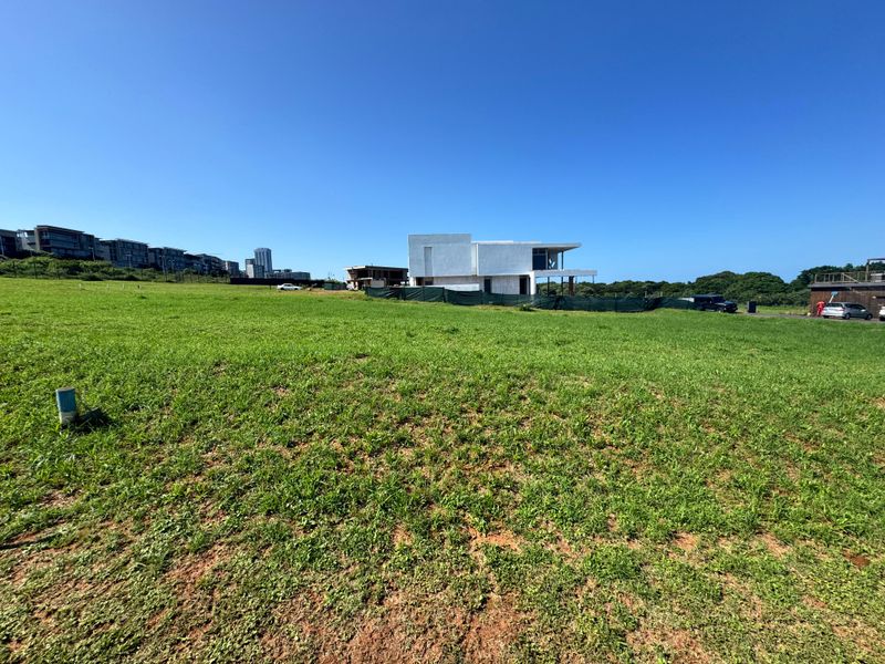 A wide-angle view of a vacant residential plot featuring open grassed land with surrounding neighborhood development visible in the background.