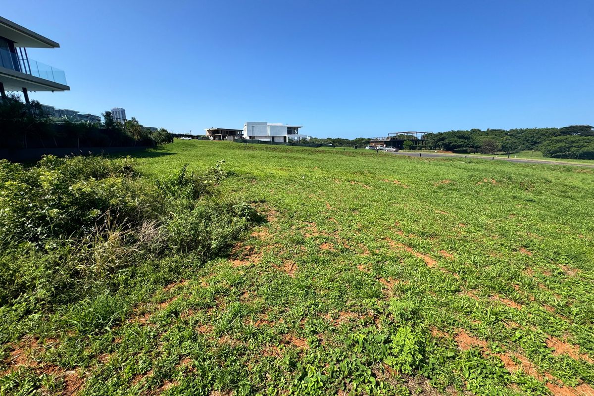 A wide-angle view of a vacant residential plot featuring open grassland with surrounding modern architectural developments in the distance.
