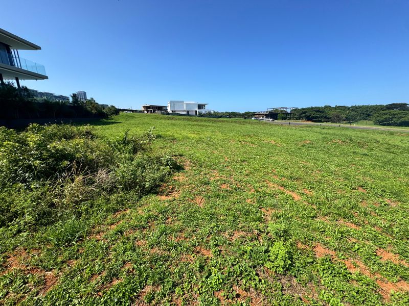 A wide-angle view of a vacant residential plot featuring open grassland with surrounding modern architectural developments in the distance.