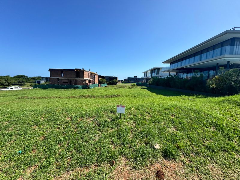 A vacant residential plot of land featuring a grass lawn, situated in a developed neighborhood with surrounding modern residential properties.