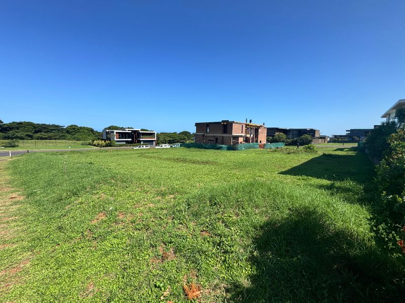 A wide-angle view of a vacant residential plot featuring open grass lawn with surrounding neighborhood development visible in the background.
