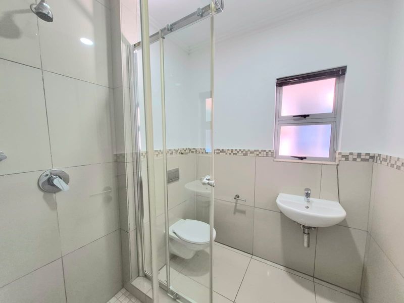A clean, modern bathroom featuring a glass-enclosed shower, wall-hung toilet, and wall-mounted basin with neutral floor-to-ceiling tiling.