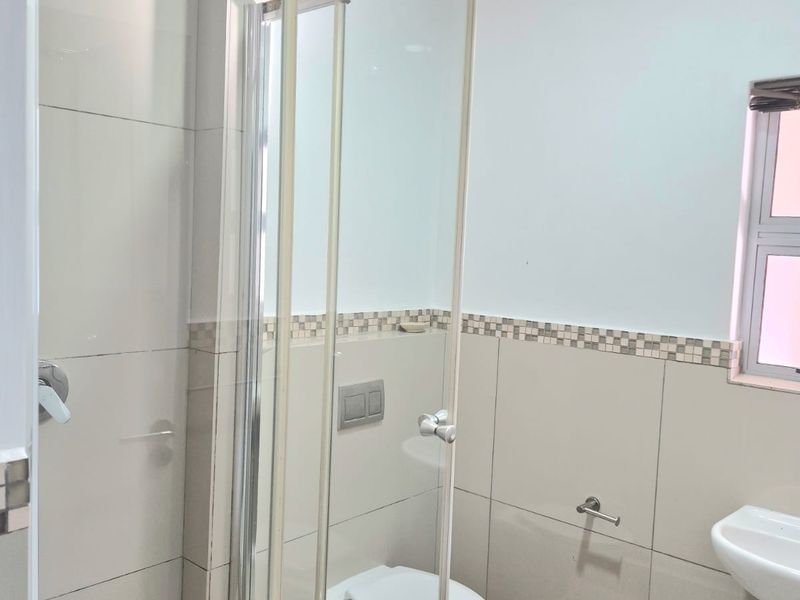 A clean, modern bathroom featuring a wall-mounted toilet, a glass-enclosed shower, and a wall-mounted basin with neutral ceramic tiling.