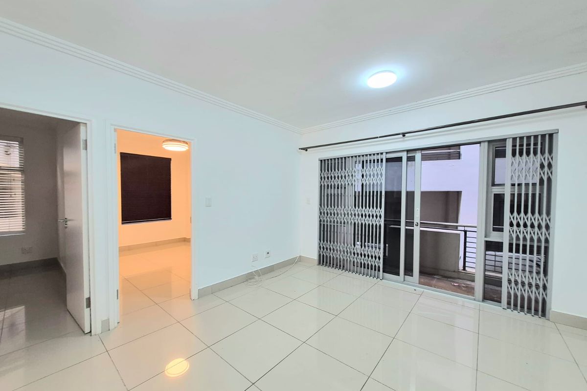 A bright, empty living area featuring neutral tiled flooring, sliding glass doors with security gates, and access to an adjoining room.